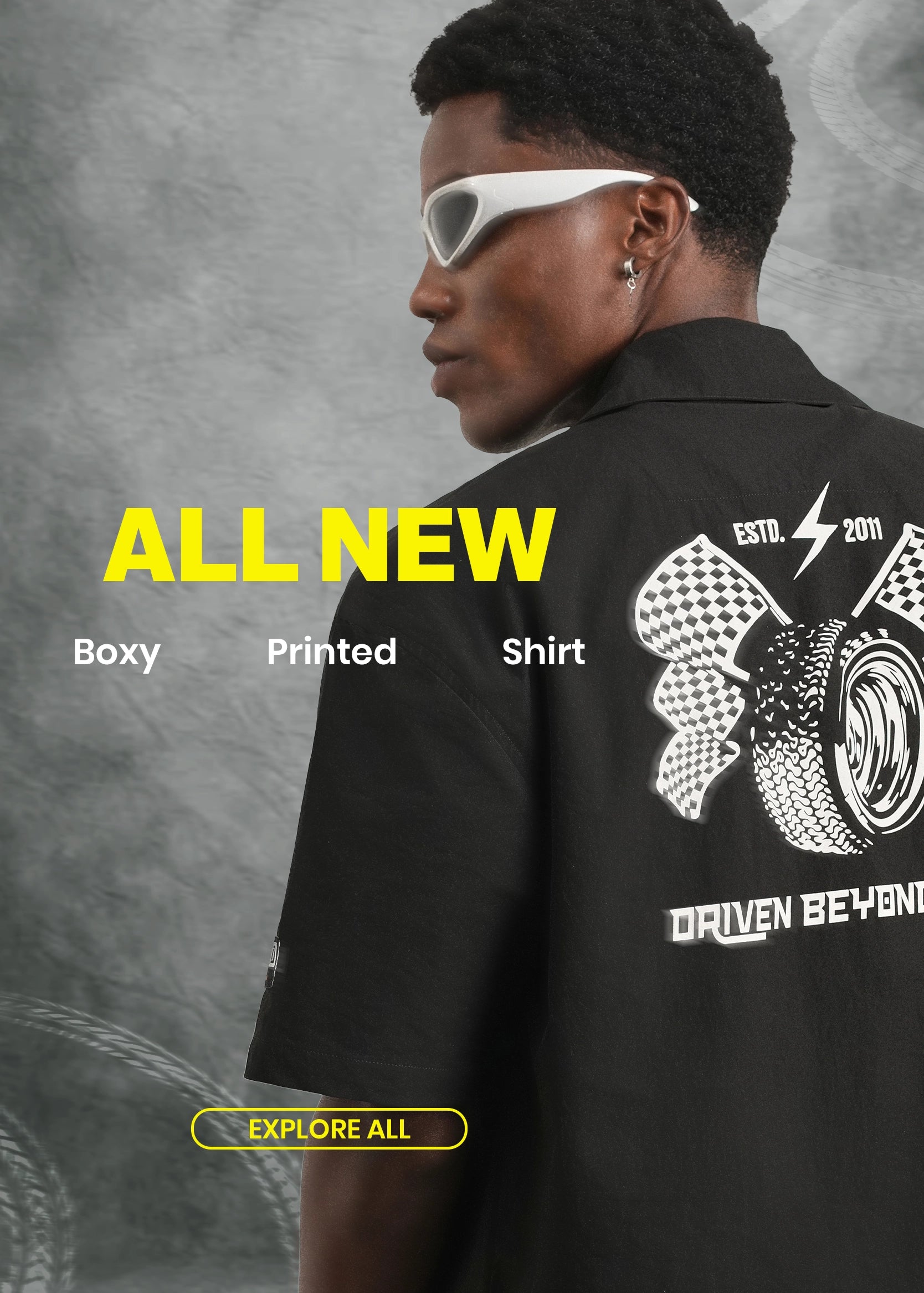 A man shown from the back wearing a black short-sleeve shirt with a large white graphic print on the back, styled with white sunglasses against a textured gray background. The image features promotional text overlays reading 'ALL NEW', 'Boxy Printed Shirt', and 'EXPLORE ALL'.