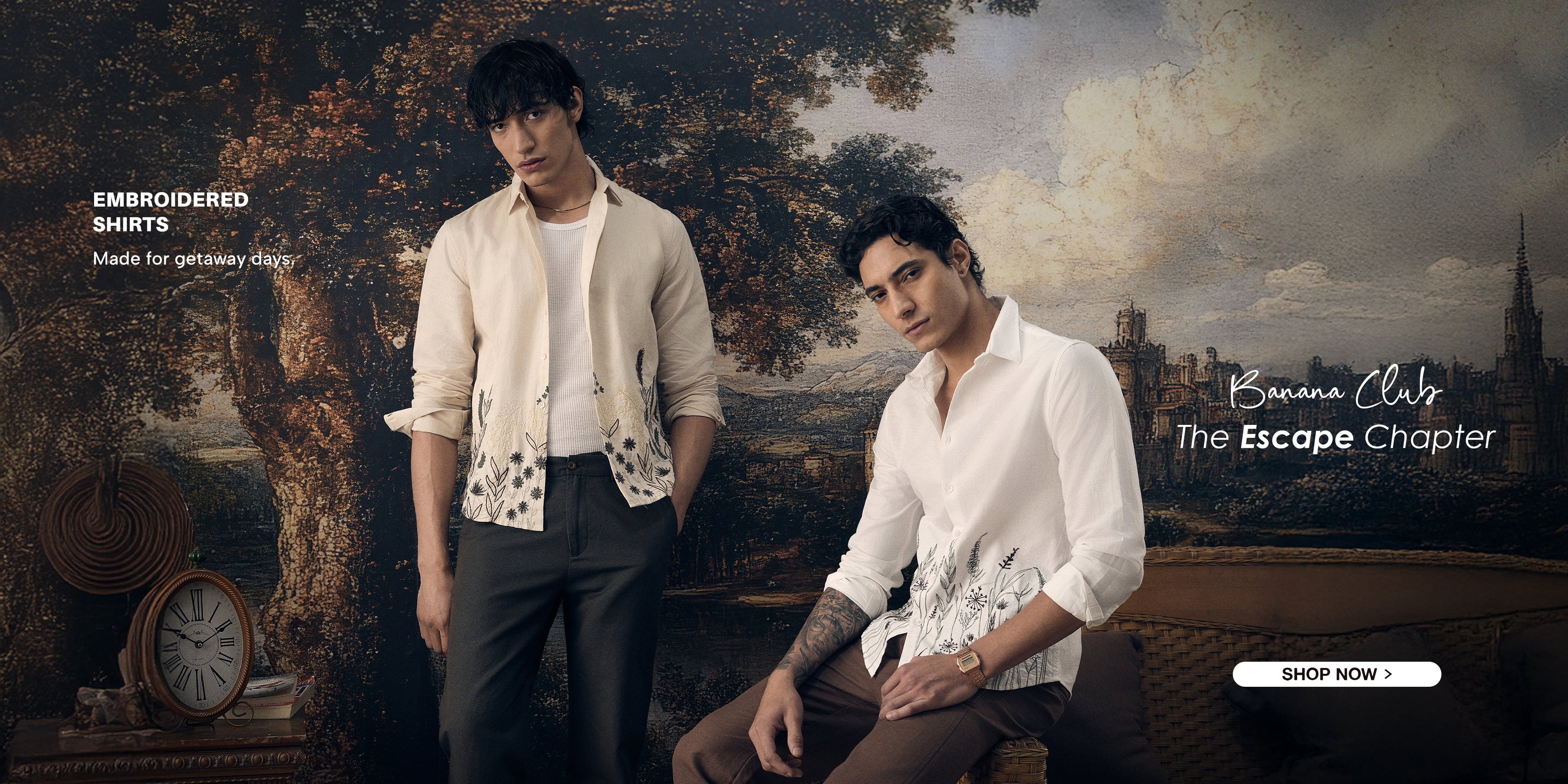 Two men in embroidered shirts standing in a scenic outdoor setting with classical painting elements.